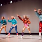 Five dancers in colorful aerobic outfits in energetic movement.