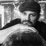 Portrait of Pekka Heikkinen. Pekka, wearing a black cap, looks at the camera and rests his chin on the back of his hand. There is a mirror ball under his palm. He has a dark beard and a black shirt.