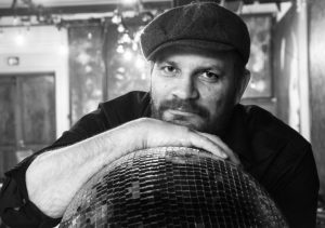 Portrait of Pekka Heikkinen. Pekka, wearing a black cap, looks at the camera and rests his chin on the back of his hand. There is a mirror ball under his palm. He has a dark beard and a black shirt.